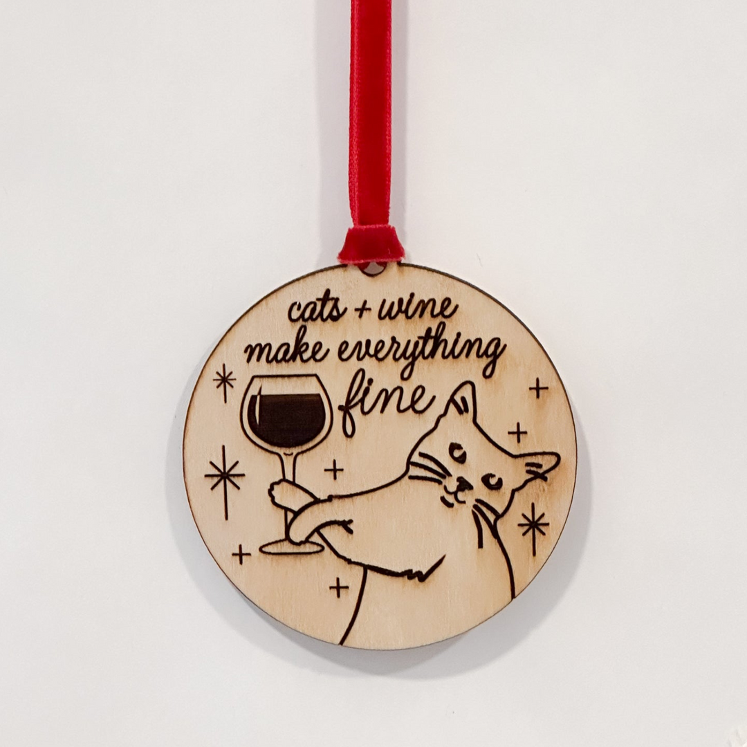 A round wooden Christmas ornament featuring a laser-engraved illustration of a cat holding a wine glass with the text 'cats + wine make everything fine', hanging from a red ribbon on a plain white background.
