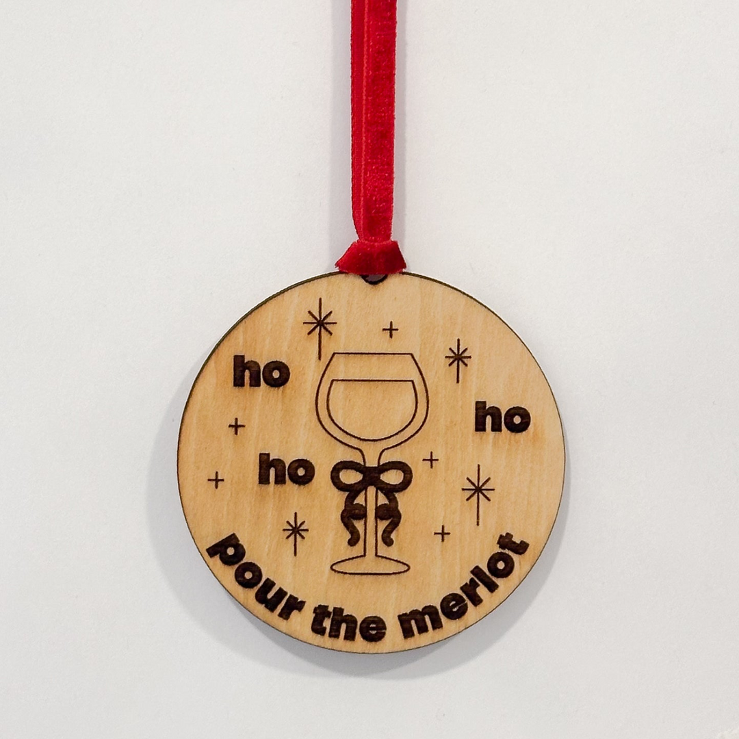 A round wooden Christmas ornament with a red hanging ribbon, featuring a wine glass illustration and the engraved text 'ho ho ho pour the merlot' on a plain white background.