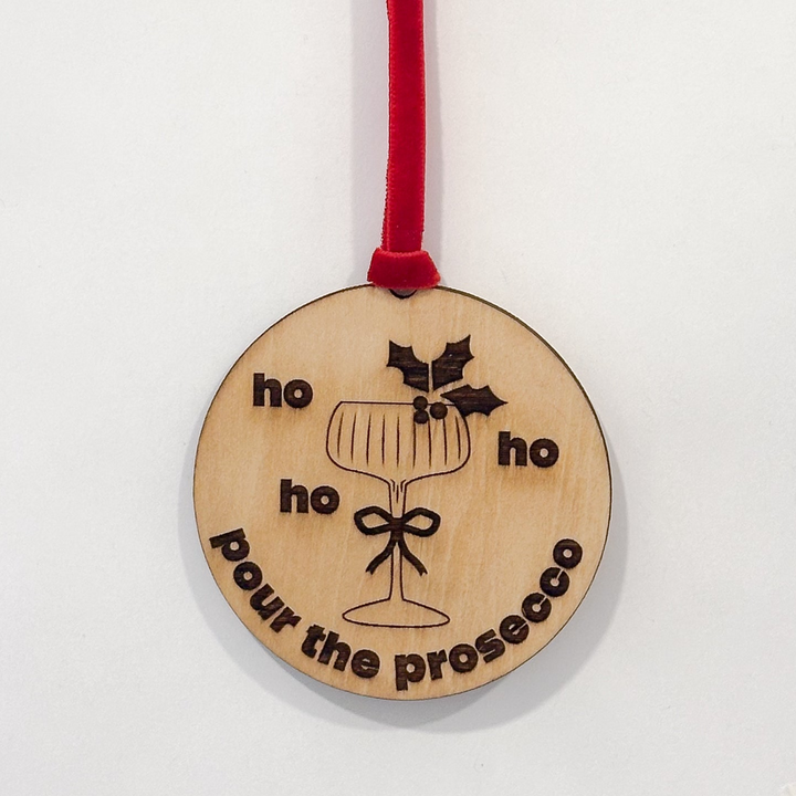A round wooden Christmas ornament with a red hanging ribbon, featuring a wine glass illustration and the text &