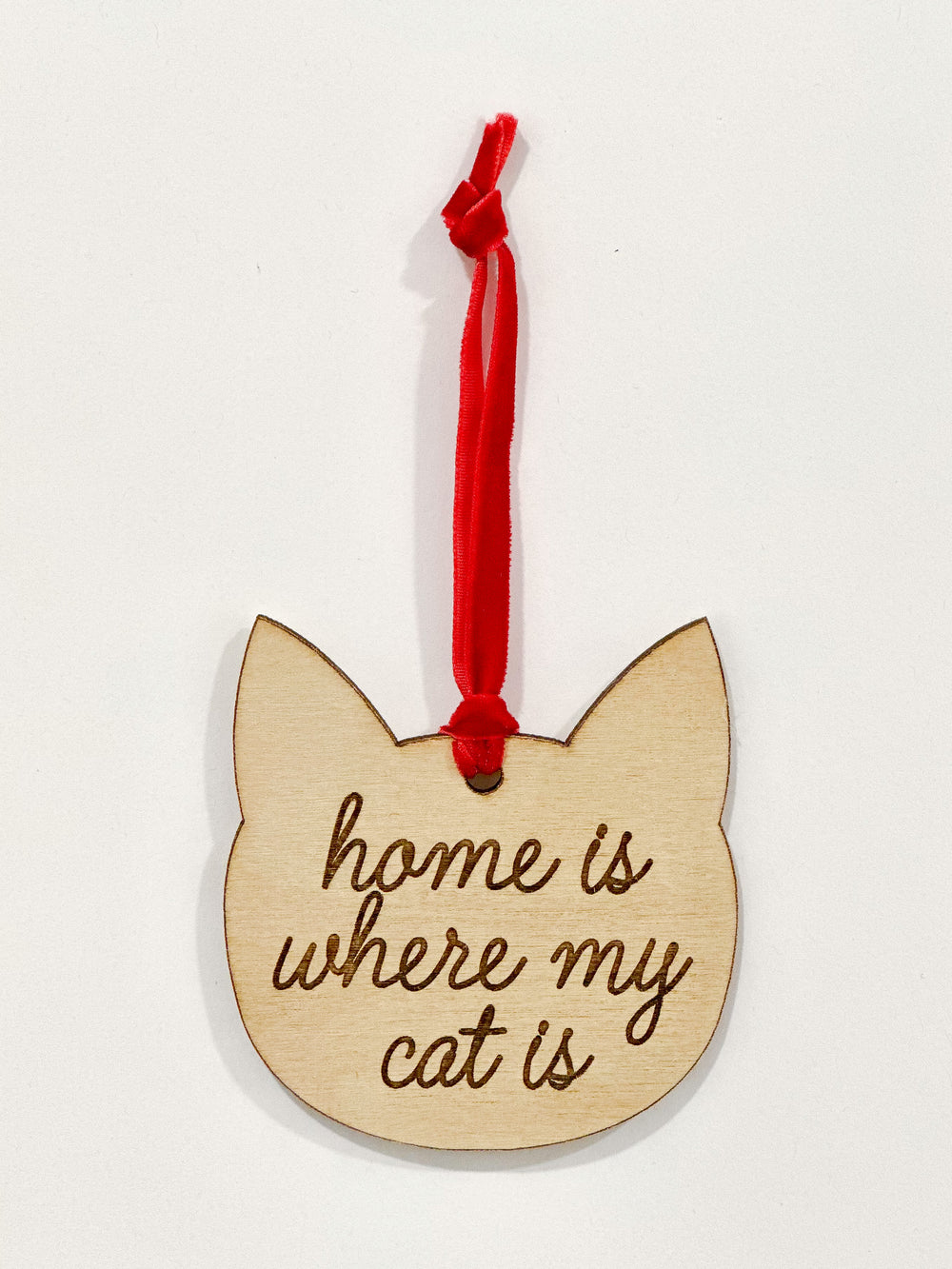 A wooden cat-shaped ornament with the engraved phrase 'home is where my cat is' in dark brown script, hanging from a red ribbon on a plain white background.