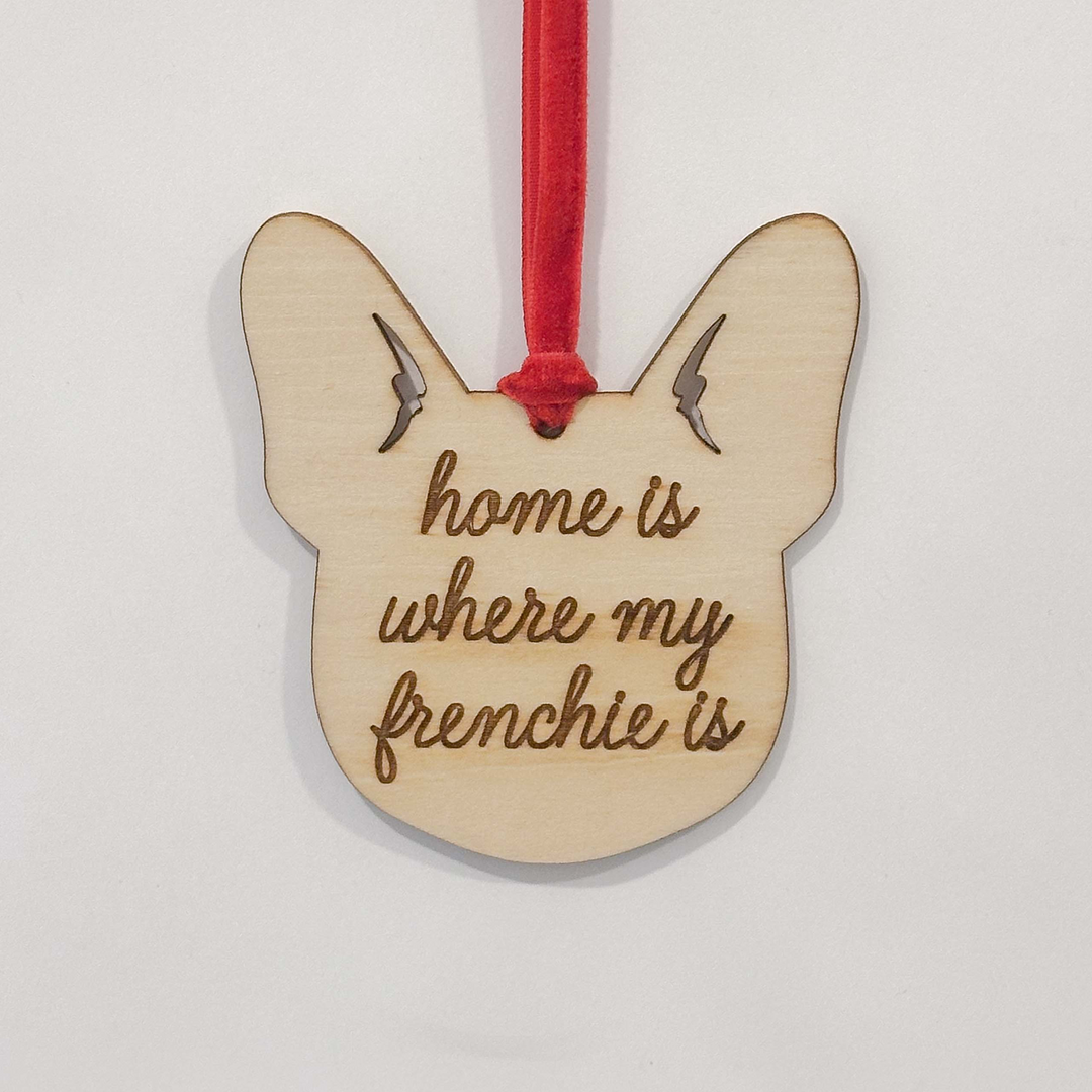 A wooden French Bulldog head hanging ornament with the engraved text 'home is where my frenchie is' and a red hanging ribbon, shown against a plain white background.