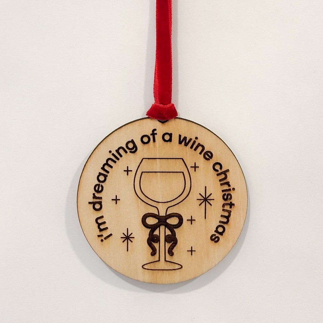 A round wooden Christmas ornament with a red hanging ribbon, featuring a laser-engraved wine glass and the text 'i'm dreaming of a wine christmas' on a plain white background.