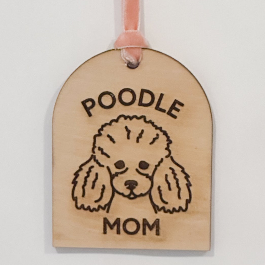 A wooden hanging ornament or tag with an arched top and a pink ribbon loop at the top. The tag is engraved with the words &
